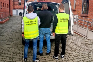  Funkcjonariusze SG w żółtych kamizelkach trzymają osobę zatrzymaną. Przed nimi stoi samochód.