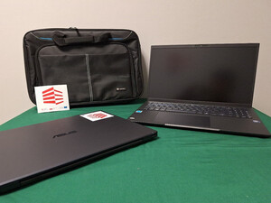 Dwa czarne laptopy. Pierwszy z lewej ma zamknięta pokrywę, za nim postawiona jest torba do przenoszenia laptopa. Z prawej kolejny laptop z otwarta pokrywą. Dwa czarne laptopy. Pierwszy z lewej ma zamknięta pokrywę, za nim postawiona jest torba do przenoszenia laptopa. Z prawej kolejny laptop z otwarta pokrywą.