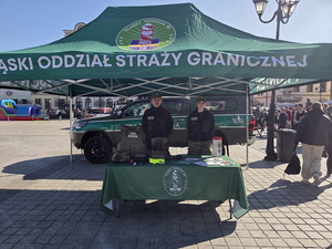 Funkcjonariusze SG stoją w namiocie za stołem na którym znajdują się materiały reklamujące Straż Graniczną. W tle samochód SG, z prawej str. spacerują ludzie. Funkcjonariusze SG stoją w namiocie za stołem na którym znajdują się materiały reklamujące Straż Graniczną. W tle samochód SG, z prawej str. spacerują ludzie.