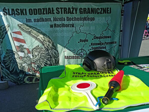 Stół a na nim żółta kamizelka SG, tarczka stop, latarka z nakładką, w tle ścianka z napisem Śląski Oddział Straży Granicznej. Stół a na nim żółta kamizelka SG, tarczka stop, latarka z nakładką, w tle ścianka z napisem Śląski Oddział Straży Granicznej.