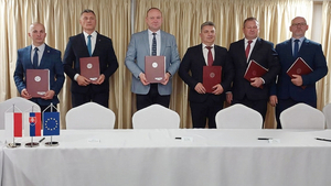  Osoby stoją za stołem, w dłoniach trzymają teczki