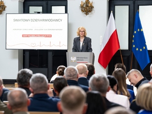  Ekran multimedialny z wyświetlonymi napisami: Światowy Dzień Krwiodawcy. Honorowy Dawca Krwi – Zasłużony dla Zdrowia Narodu. Łączy nas krew, która ratuje życie. Przy mównicy Pani Minister Urszuli Demkow. Z Jej lewej strony flaga Polski i Unii Europejskiej.
