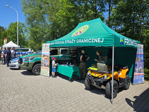  Samochód Straży Granicznej, obok namiot. Wewnątrz stoją funkcjonariusze, na stole materiały promocyjne. Przed stołem mały chłopiec. W namiocie z prawej strony postawiony quad.