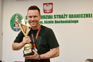  Radosny zawodnik drużyny zwycięskiej. Prawa dłoń ma zaciśnięta, w lewej trzyma puchar za pierwsze miejsce. Za nim na ścianie napis: Śląski Oddział Straży Granicznej im. nadkom. Józefa Bocheńskiego w Raciborza.