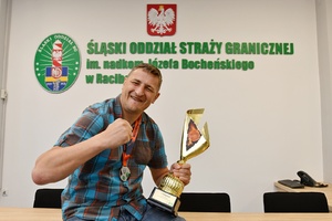  Radosny zawodnik drużyny zwycięskiej. Prawa dłoń ma zaciśnięta, w lewej trzyma puchar za pierwsze miejsce. Za nim na ścianie napis: Śląski Oddział Straży Granicznej im. nadkom. Józefa Bocheńskiego w Raciborza.