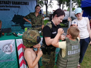 Stoisko wystawiennicze Straży Granicznej. Funkcjonariuszka pomaga ubrać chłopcu kamizelkę. 2 Chłopiec ma ubrany już hełm.