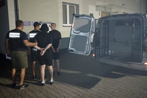  Samochód do przewozu osób zatrzymanych, otwarte tylne drzwi. Dwóch funkcjonariuszy prowadzi dwie osoby zatrzymane