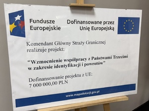 Tablica z napisami: Komendant Główny Straży Granicznej realizuje projekt: "Wzmocnienie współpracy z Państwami Trzecimi w zakresie identyfikacji i powrotów". Dofinansowanie projektu z UE: 7 000 000,00 PLN. Fundusze Europejskie. Dofinansowane przez Unię Europejską.