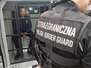  Funkcjonariusz Straży Granicznej stoi tyłem, na kamizelce taktycznej napis: Straż Graniczna Polish Border Guard. Osoba zatrzymana siedzi za kratą w samochodzie do przewozu osób zatrzymanych.