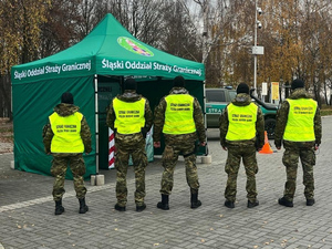 5 funkcjonariuszy Straży Granicznej w żółtych kamizelkach. Za nimi namiot SG i samochów