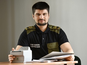 Funkcjonariusz Straży Granicznej. Siedzi przy stole na którym stoi maszyna do pobierania odcisków palców i teczki z dokumentami.