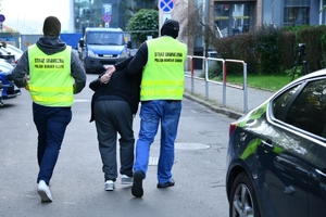 Funkcjonariusze w żółtych kamizelkach z napisem Straż Graniczna prowadza osobę. Ma ona założone kajdanki.